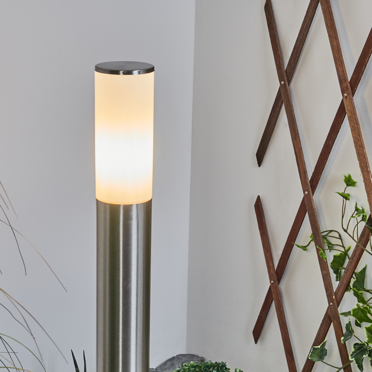 Eine zylindrische Außenlampe leuchtet neben einem Holzgitter mit Efeu. Die Lampe ist silberfarben und beleuchtet eine weiße Wand.