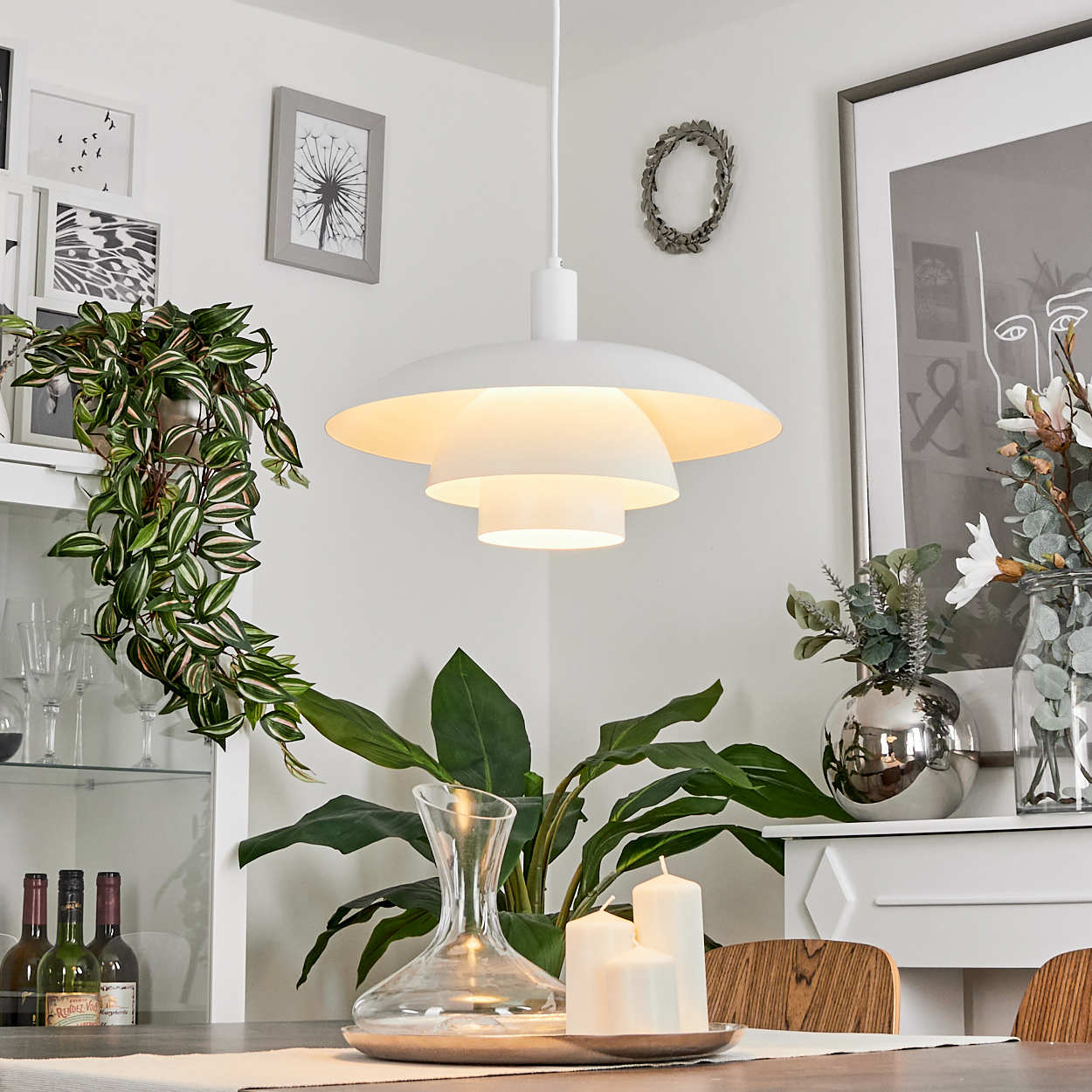 Ein Esszimmer mit weißer Lampe, Pflanzen, Kerzen und Kunstwerken. Eine Glaskaraffe und eine silberne Vase sind ebenfalls vorhanden.