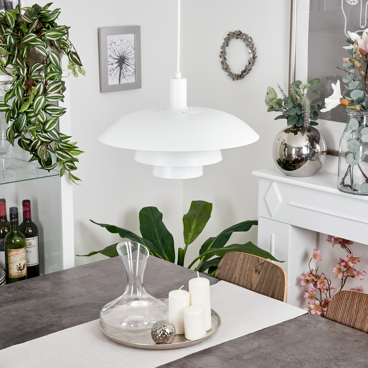 Eine weiße, mehrschichtige Pendelleuchte hängt über einem grauen Tisch mit Kerzen und einer Karaffe. Pflanzen und Blumen sind im Hintergrund.