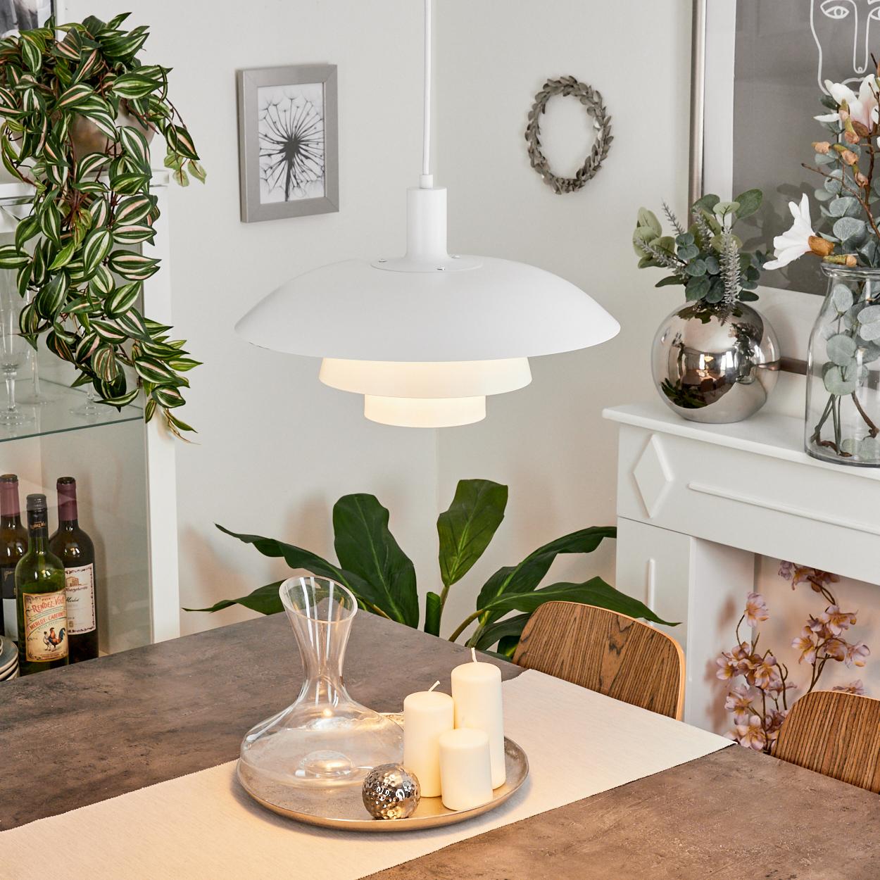 Esszimmer mit weißer Lampe, Tischdeko, Pflanzen und Dekor. Kerzen, Wein und silbernes Ornament sind zu sehen.