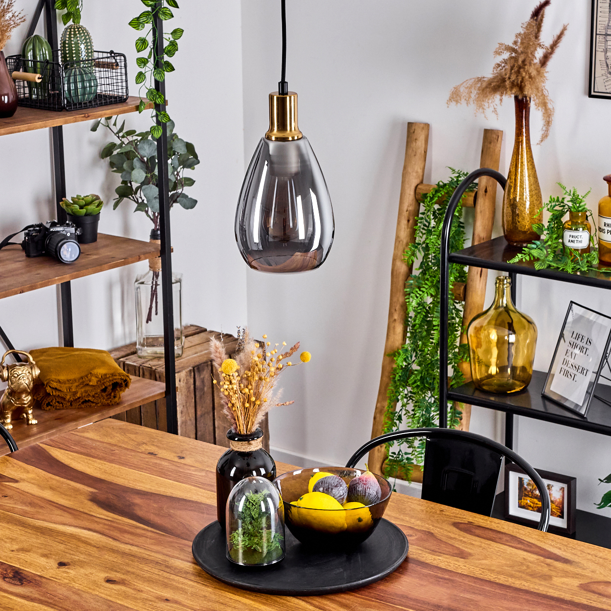 Ein Raum mit Holztisch und Regalen. Eine Hängelampe, Früchte, Vasen und Pflanzen stehen auf dem Tisch und den Regalen.