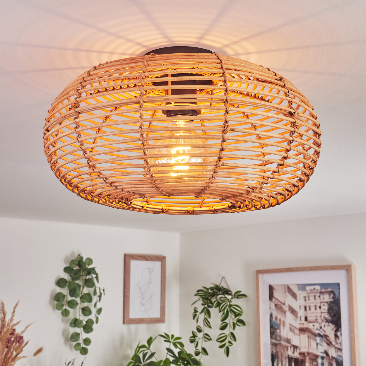 Eine gewebte Rattanlampe hängt an einer weißen Decke. Darunter sind gerahmte Kunstwerke und hängende Pflanzen zu sehen.