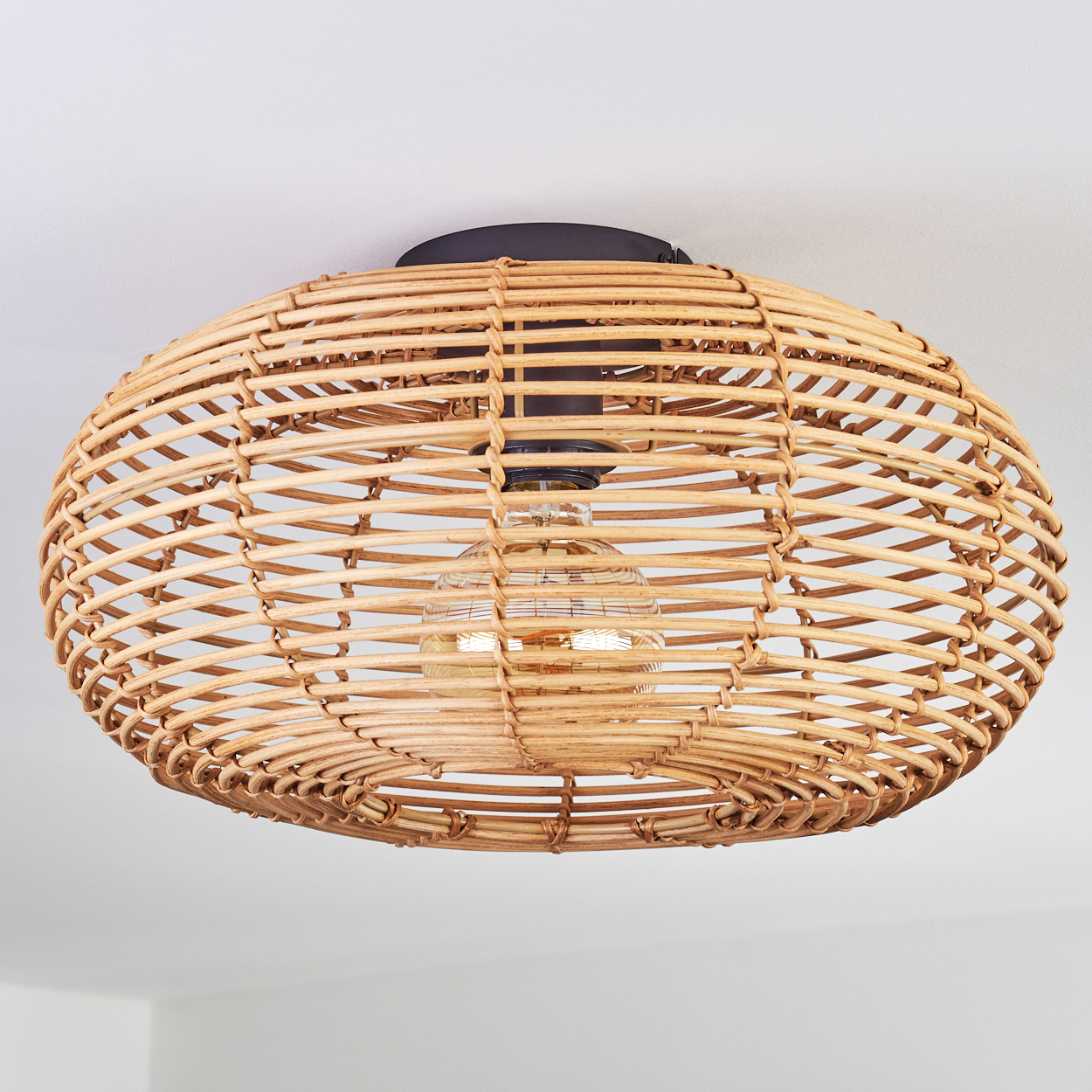 Eine runde Pendelleuchte aus Rattan mit sichtbarer Glühbirne. Das Licht hängt an einer weißen Decke. Das Licht hat eine dunkle Halterung.