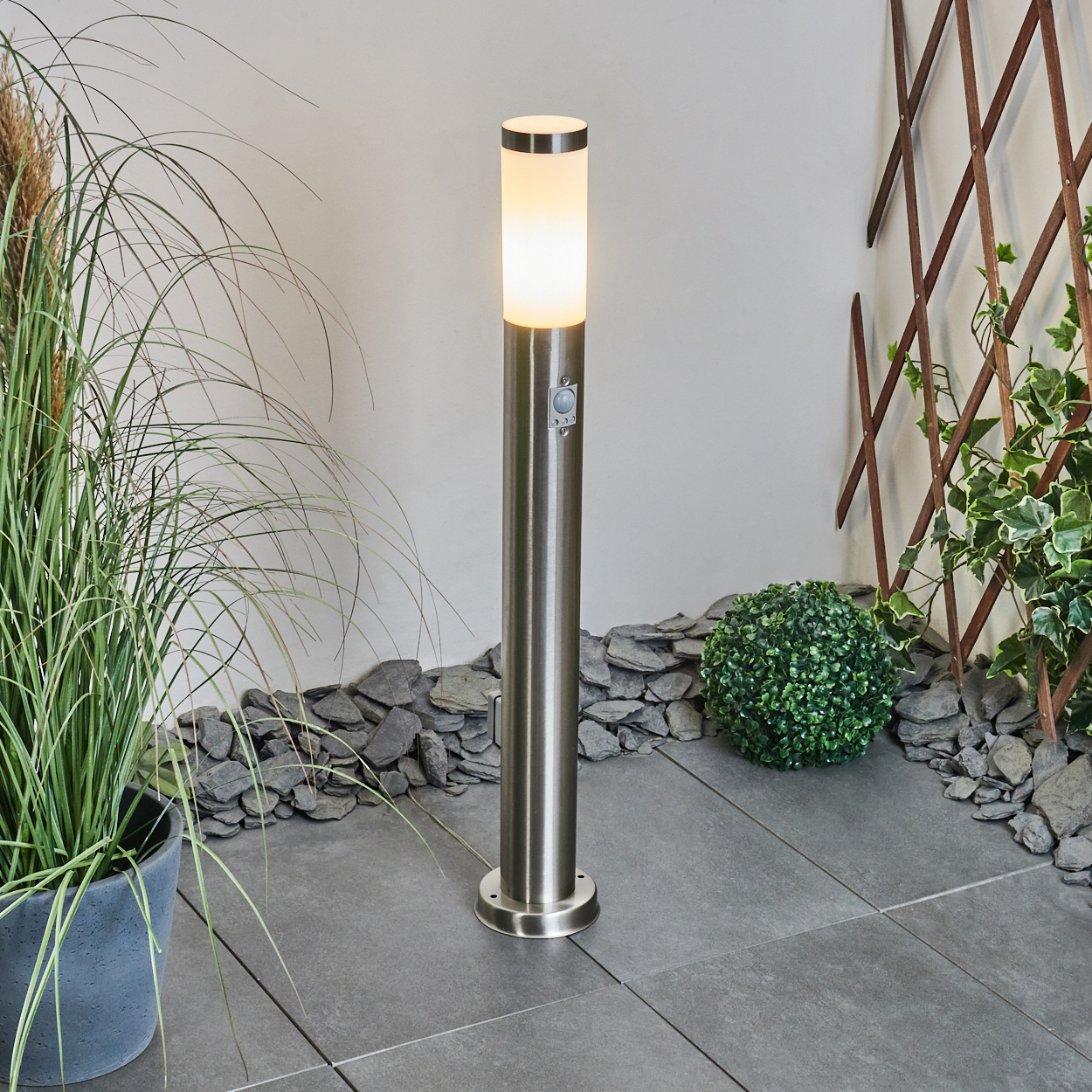 Eine beleuchtete, metallische Außenlampe steht auf grauen Fliesen. Grüne Pflanzen und ein Gitter umgeben sie. Hintergrund: weiße Wand und dunkle Steine.
