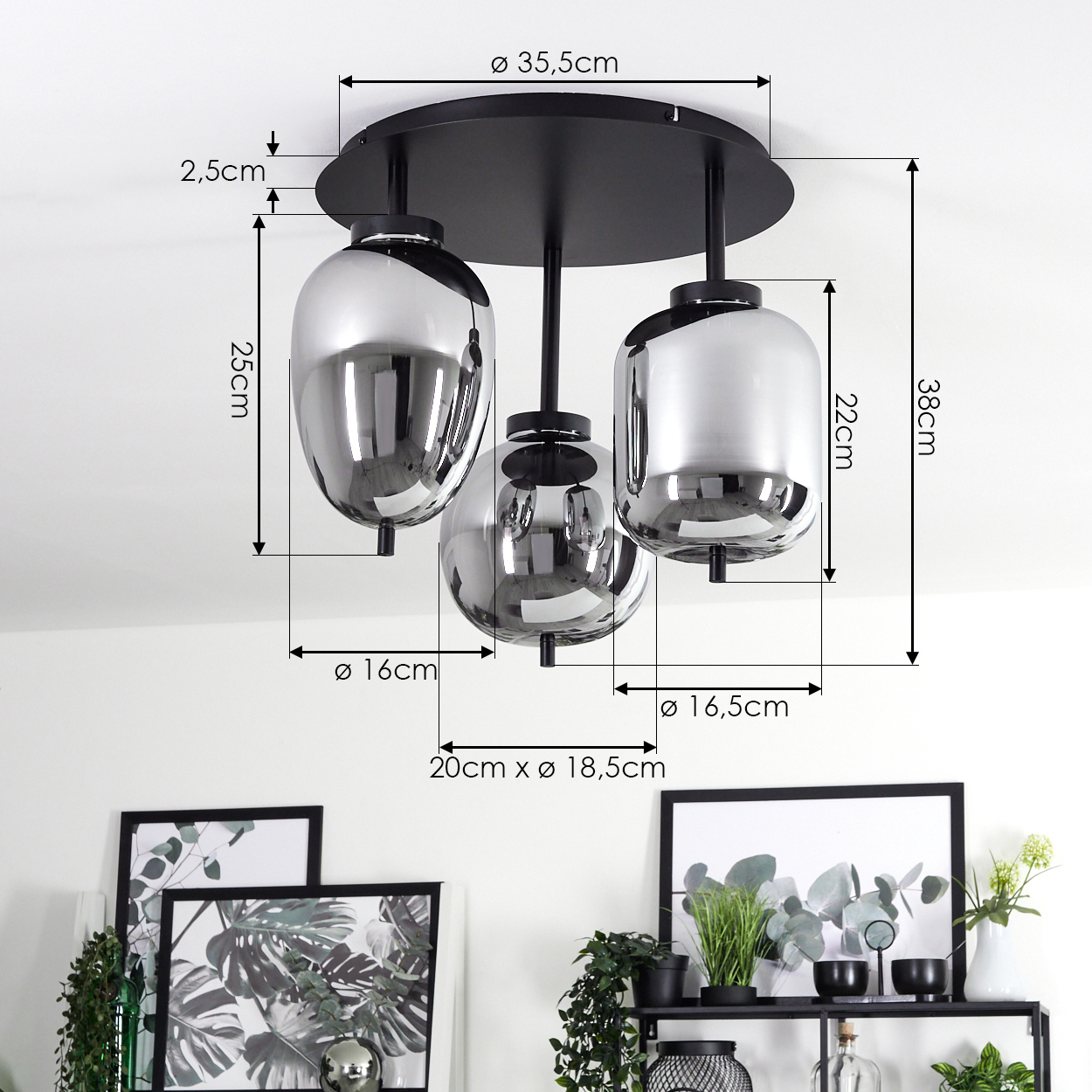 Eine Deckenleuchte mit drei glänzenden Glasschirmen. Maße sind angegeben. Darunter ein Regal mit Pflanzen und gerahmten Bildern.