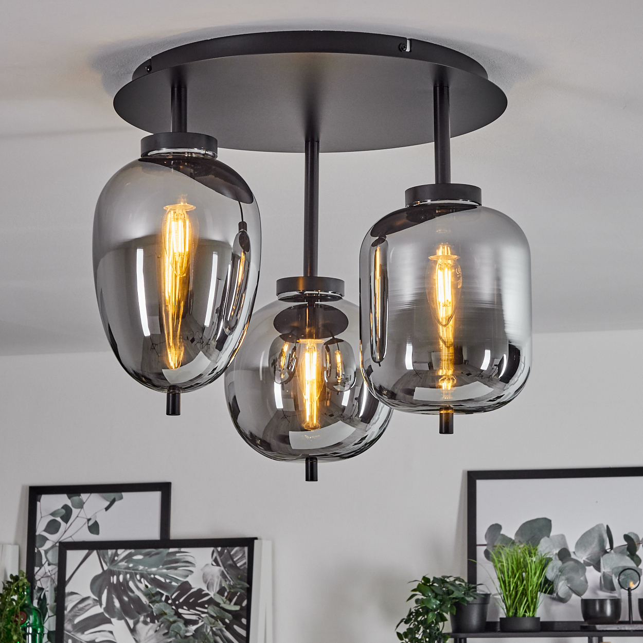 Deckenleuchte mit drei rauchfarbenen Glasschirmen, je mit sichtbarer Glühbirne. Schwarze Armaturen und weiße Decke. Deko im Hintergrund.