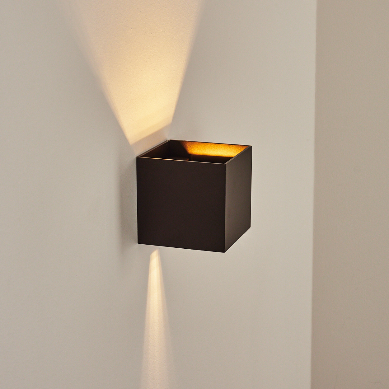 Eine dunkle, quadratische Wandleuchte strahlt nach oben und unten Licht aus und wirft Schatten auf die beige Wand. Das Innere der Lampe leuchtet.