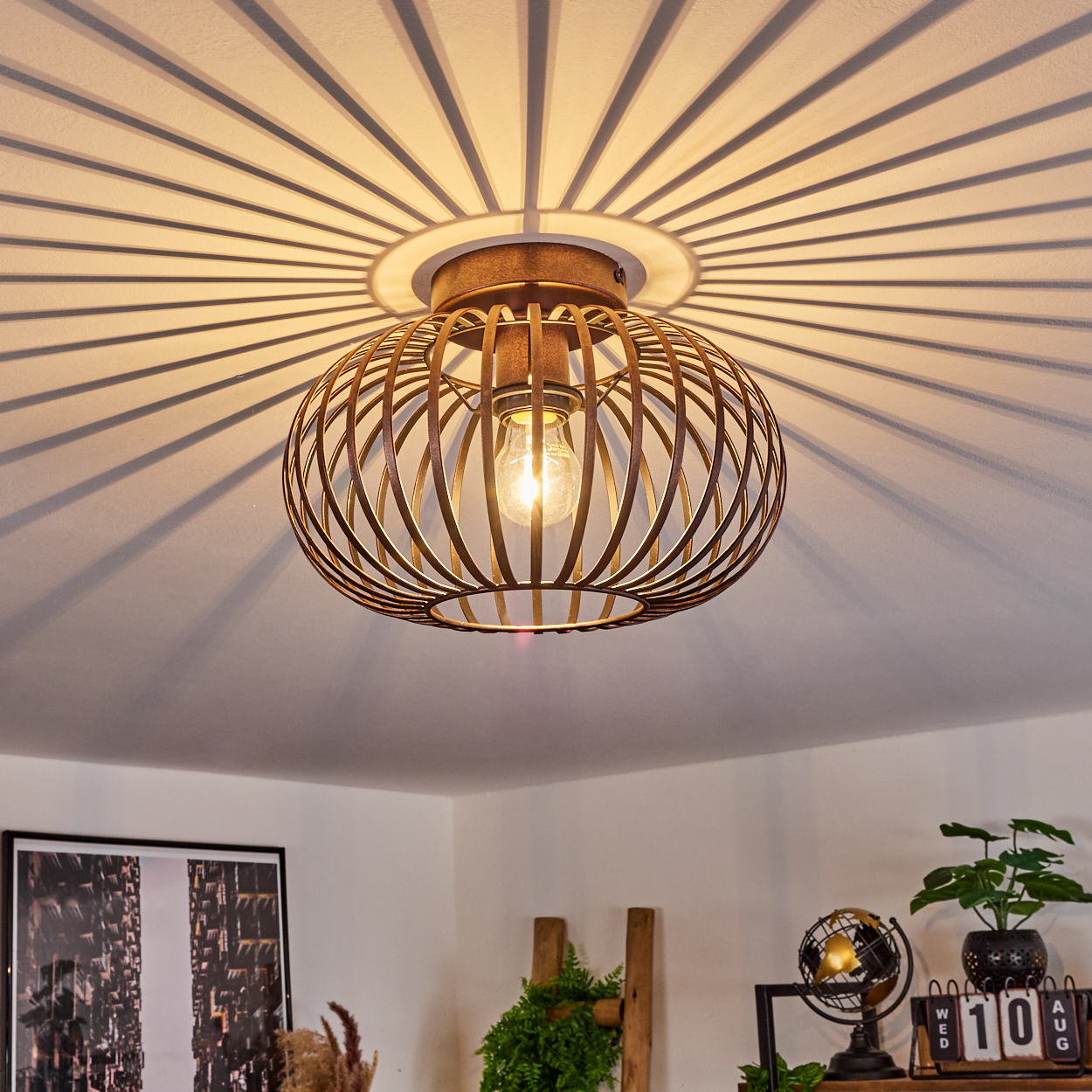 Eine bronze Cage-Deckenlampe mit einer leuchtenden Glühbirne. Die Decke wirft einen radialen Schatten. Im Hintergrund sind Dekorationsartikel.