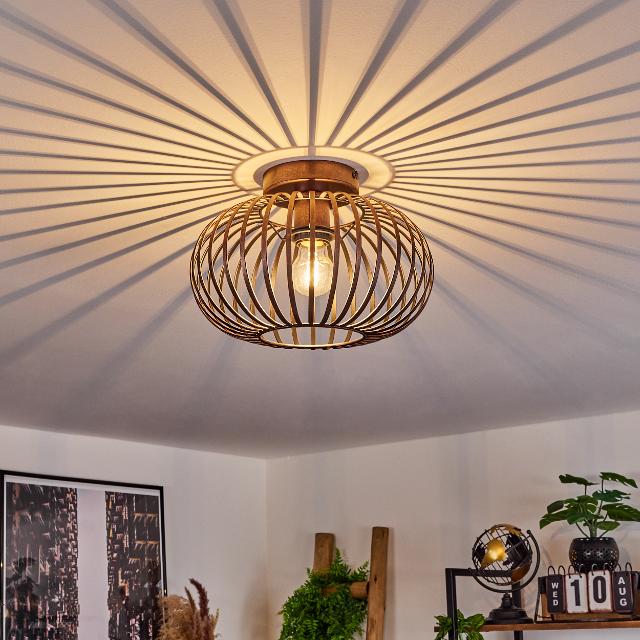 Eine Deckenleuchte mit käfigartigem Design, die radiale Schatten auf die Decke wirft. Der Raum umfasst Dekoration und Pflanzen.