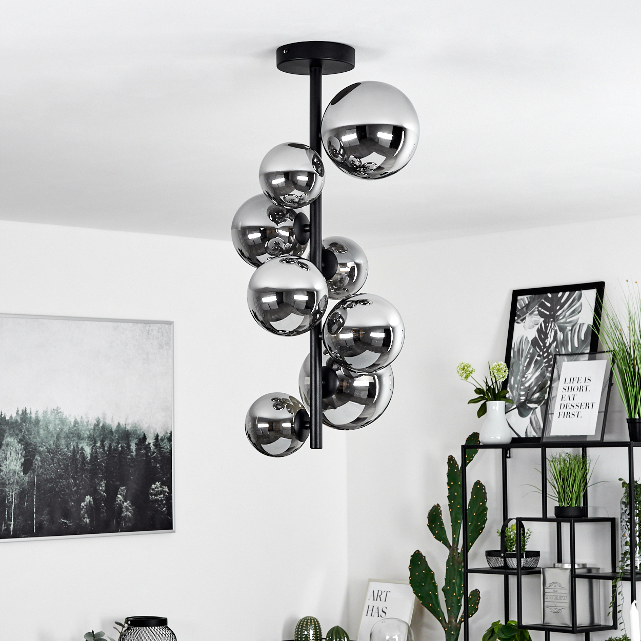 Eine moderne Deckenleuchte mit silbernen Kugeln hängt von der Decke. Ein Regal zeigt Pflanzen, Kunst und Dekorationsartikel. Weiße Wände.