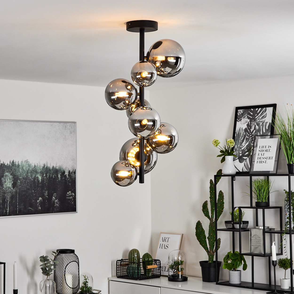 Eine moderne Deckenlampe mit Glaskugeln hängt in einem Raum mit Kunst und Pflanzen. Ein schwarzes Metallregal trägt Dekoration.