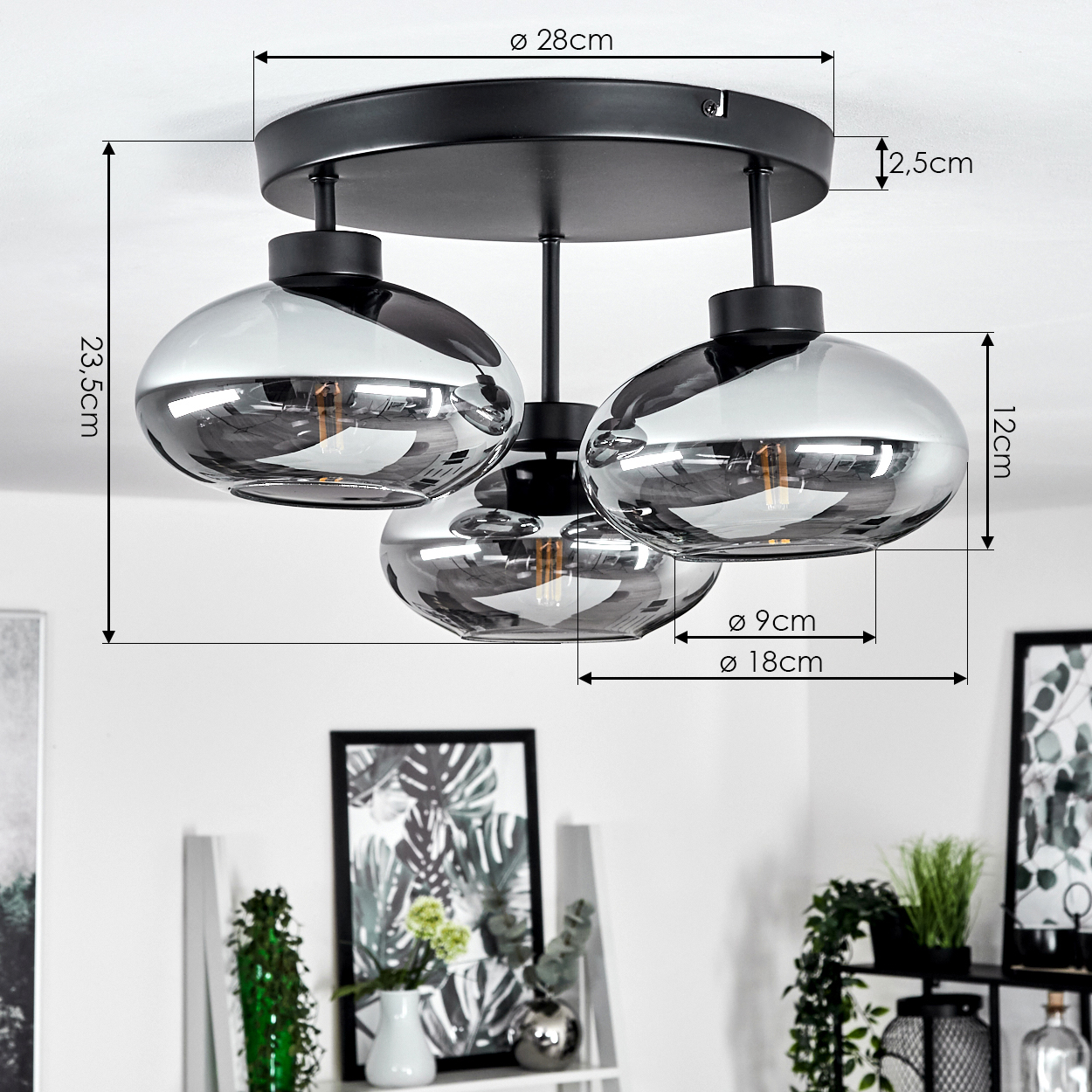 Eine schwarze Deckenlampe mit drei Glasschirmen. Abmessungen sind beschriftet. Hintergrund zeigt Deko, Pflanzen und eine weiße Wand.