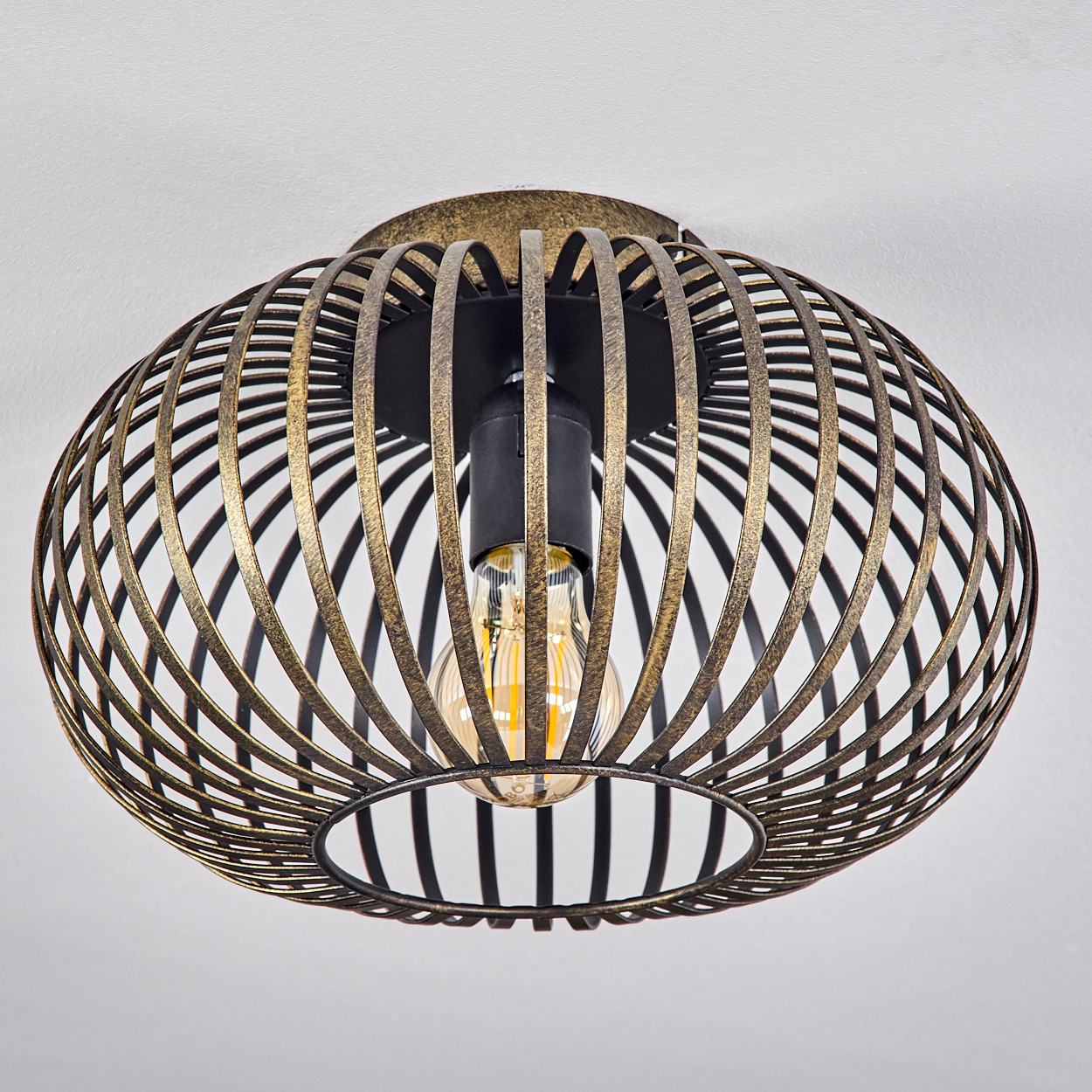 Eine moderne Deckenlampe mit einem käfigartigen Schirm in Gold und Schwarz, die eine Glühbirne beherbergt, vor einer weißen Decke.