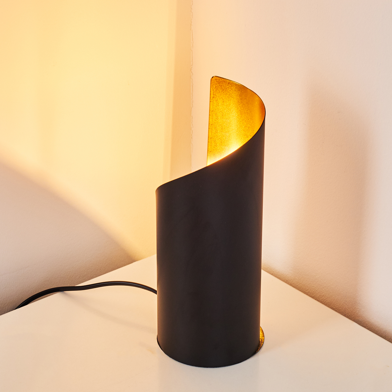 Eine schwarze zylindrische Lampe mit goldenem Innenraum steht auf einer weißen Oberfläche. Die Wand ist weiß, und Licht scheint aus der Lampe.