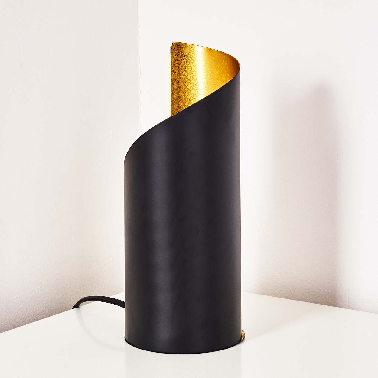 Eine schwarze zylindrische Lampe mit goldfarbenem Innenraum steht auf einer weißen Oberfläche. Der Hintergrund ist eine weiße Wand.