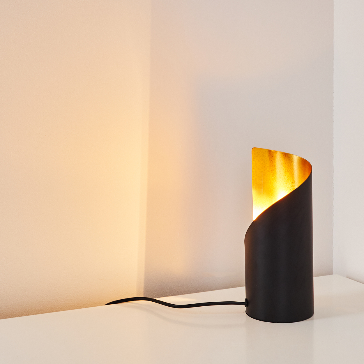 Eine schwarze zylindrische Lampe mit goldenem Innenraum steht auf einer weißen Oberfläche. Sie steht in der Ecke zweier Wände. Ein schwarzes Kabel ist befestigt.