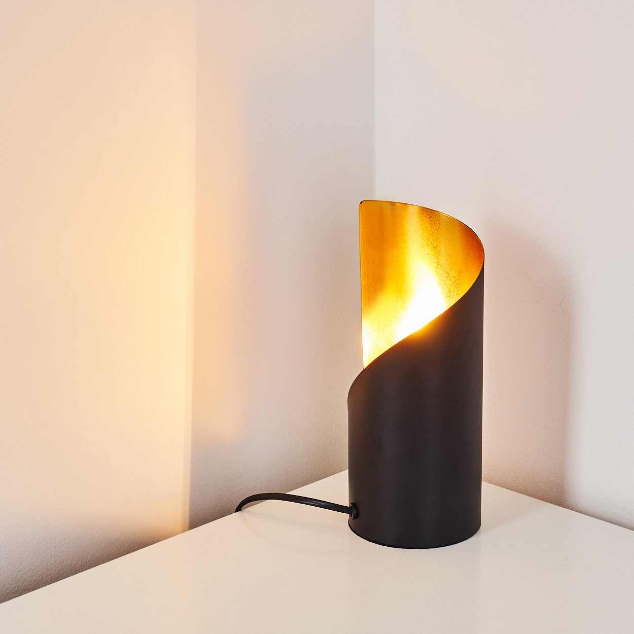 Eine schwarz-goldene Lampe auf einem weißen Tisch mit Kabel. Der Hintergrund sind weiße Wände und Licht von der Lampe.