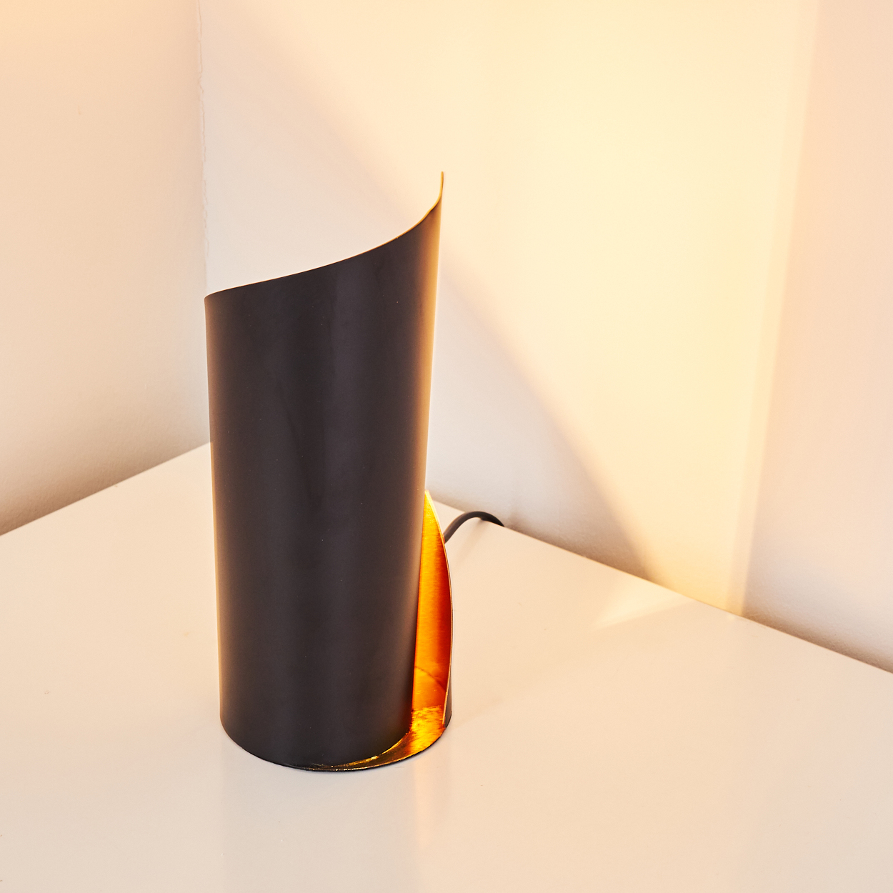 Eine moderne, schwarz-weiße, zylindrische Lampe mit goldenem Innenraum steht auf einem weißen Tisch. Der Hintergrund ist eine helle Wand.