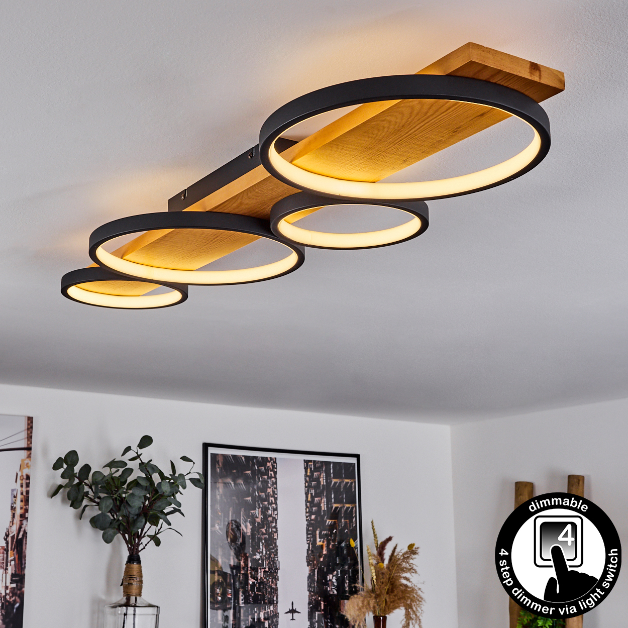 Deckenleuchte mit vier kreisförmigen Lichtern. Holzbalken und schwarze Ringe. Über Lichtschalter dimmbar. Hintergrund zeigt Kunst und Pflanzen.