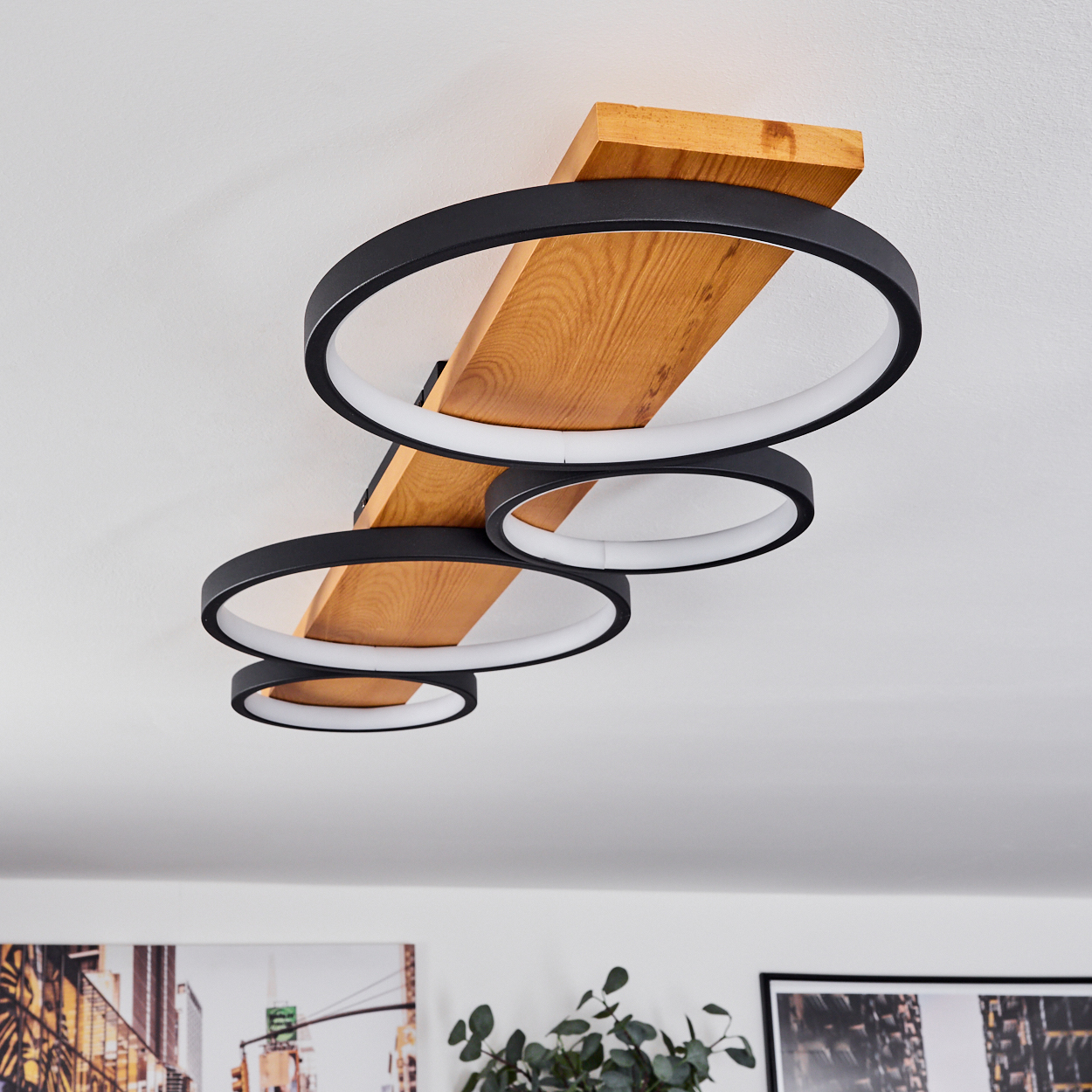 Moderne Deckenleuchte mit schwarzen Ringen und Holz. Kreise sind entlang eines Holzbalkens angeordnet. Weiße Decke im Hintergrund.