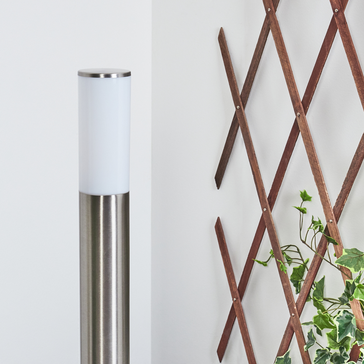 Eine silberne Lampe mit weißem Oberteil steht neben einem hölzernen Spalier mit Efeu an einer weißen Wand.