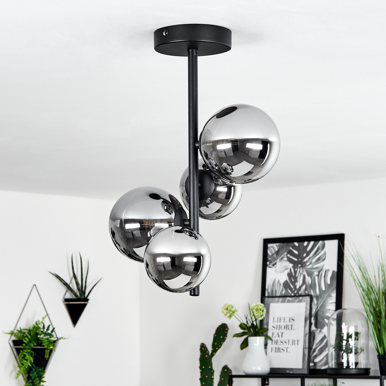 Eine moderne Deckenlampe mit silbernen Kugeln. Der Raum hat weiße Wände, ein schwarzes Regal mit einem Bild und grüne Pflanzen.