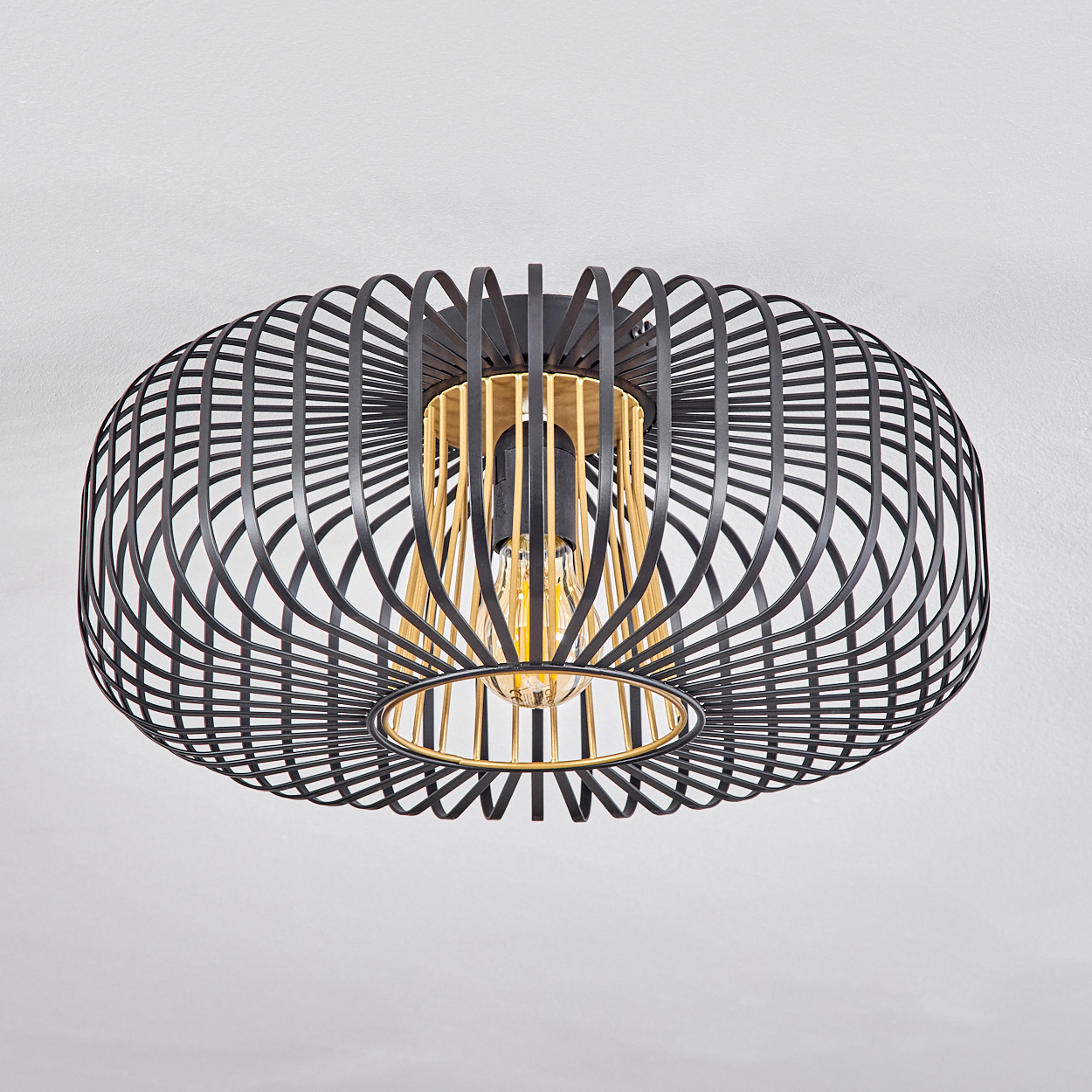 Schwarz-goldene, käfigartige Deckenlampe mit Glühbirne. Der Hintergrund ist eine schlichte, weiße Decke.