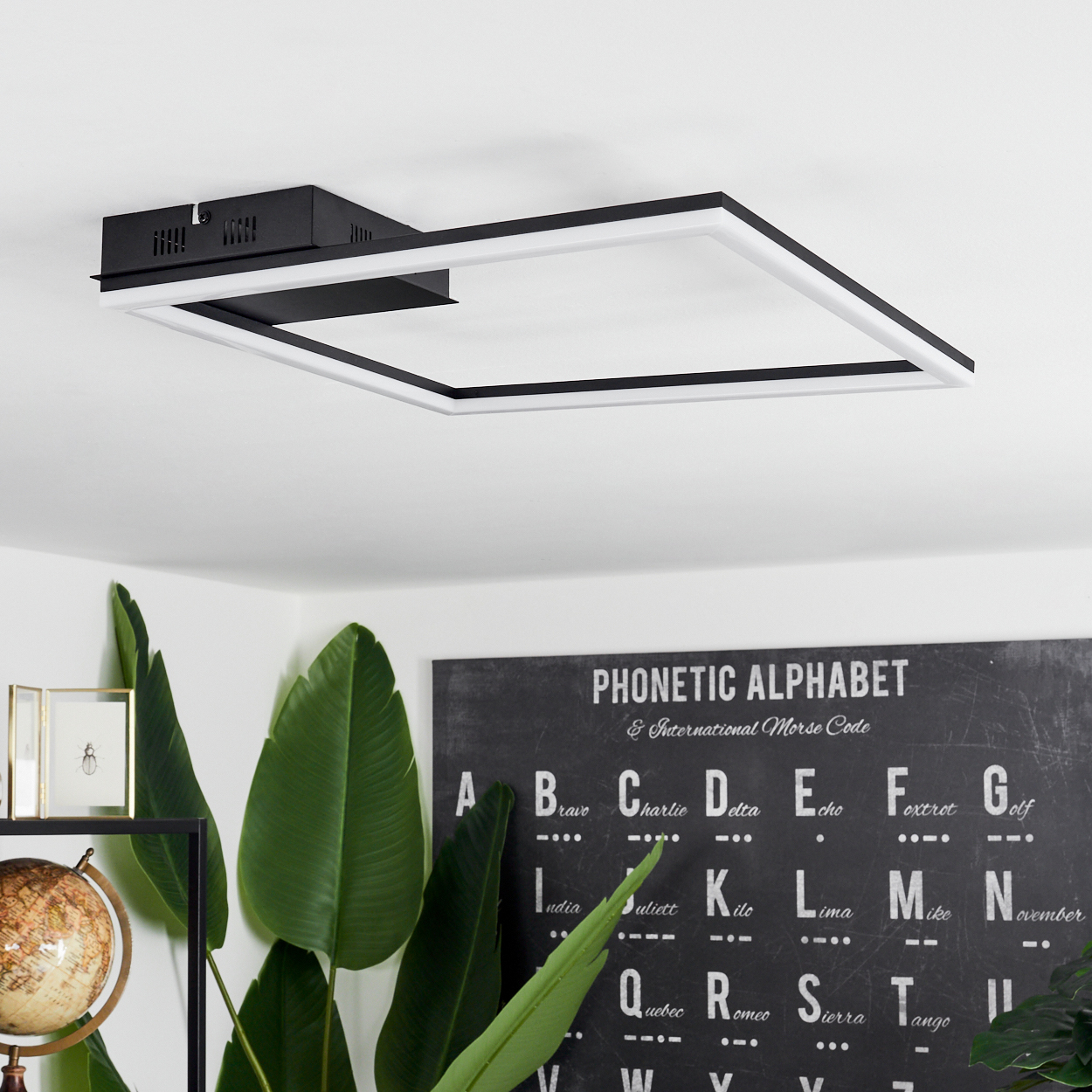 Eine schwarz-weiße quadratische Deckenlampe. Darunter ein phonetisches Alphabet-Poster, grüne Blätter, ein Globus und ein Bilderrahmen.