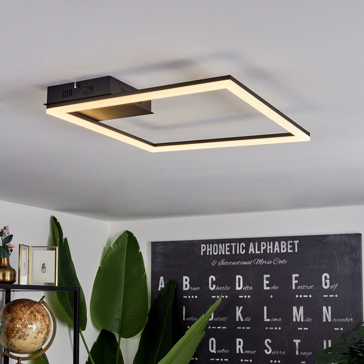 Eine quadratische Deckenleuchte beleuchtet einen Raum. Darunter stehen Pflanzen und ein Globus neben einer Wand mit einem phonetischen Alphabet.