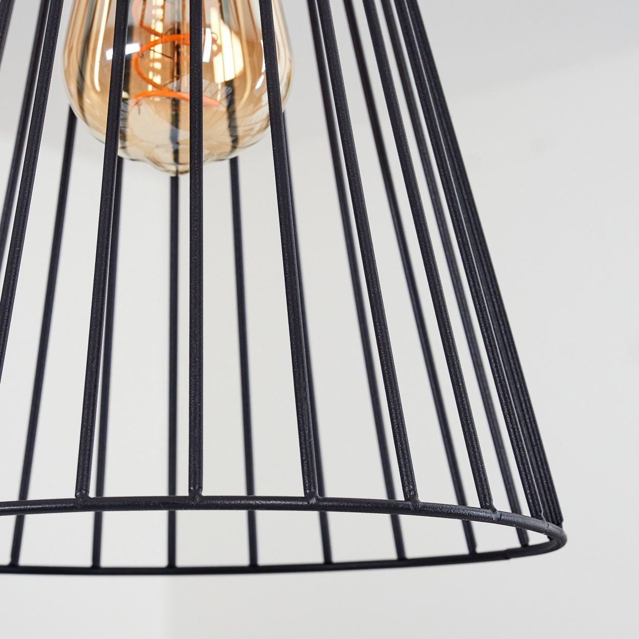 Ein schwarzer, käfigartiger Lampenschirm umschließt eine Glühbirne. Die Glühbirne ist beleuchtet, und der Hintergrund ist weiß. Die Lampe hängt von oben.