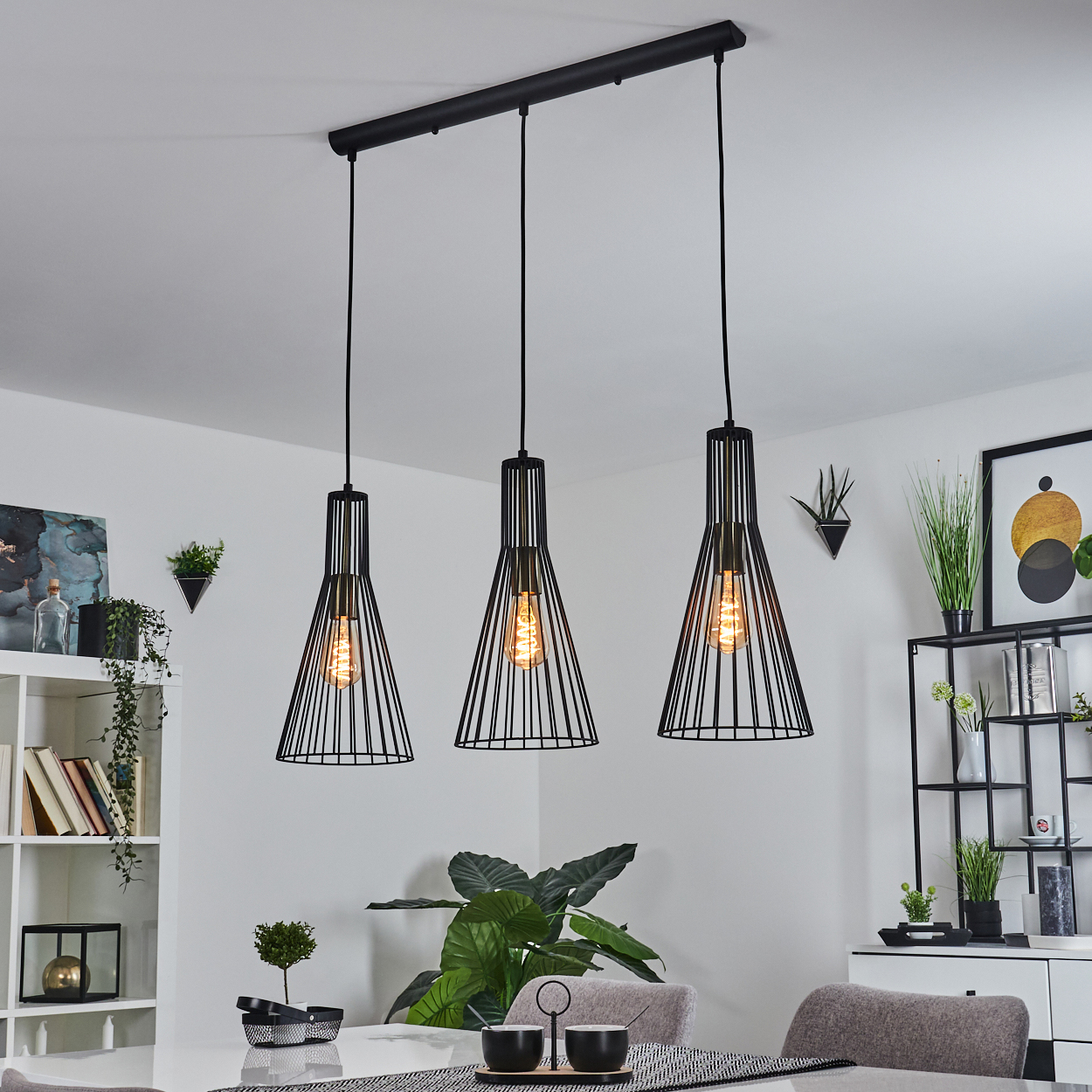 Drei schwarze Pendelleuchten mit freiliegenden Glühbirnen hängen über einem weißen Tisch. Pflanzen und Dekoration befinden sich im Hintergrund, mit weißen Wänden.