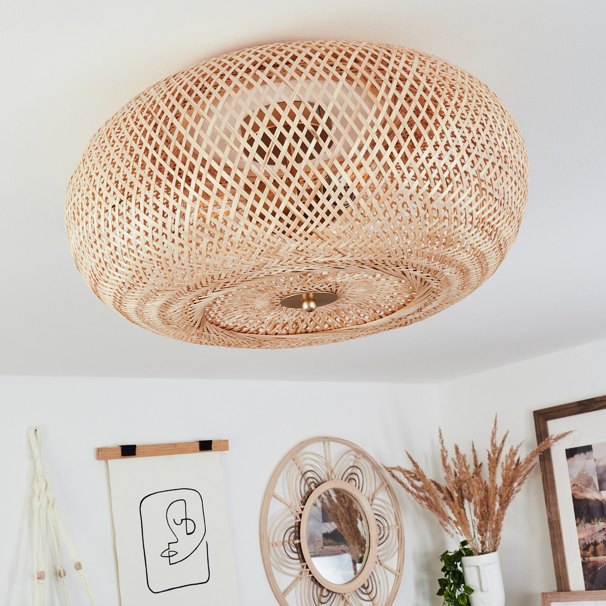 Gewebte Deckenlampe aus Rattan. Darunter eine Wand mit Kunst, Spiegel, Vase mit getrocknetem Gras und einem gerahmten Bild.