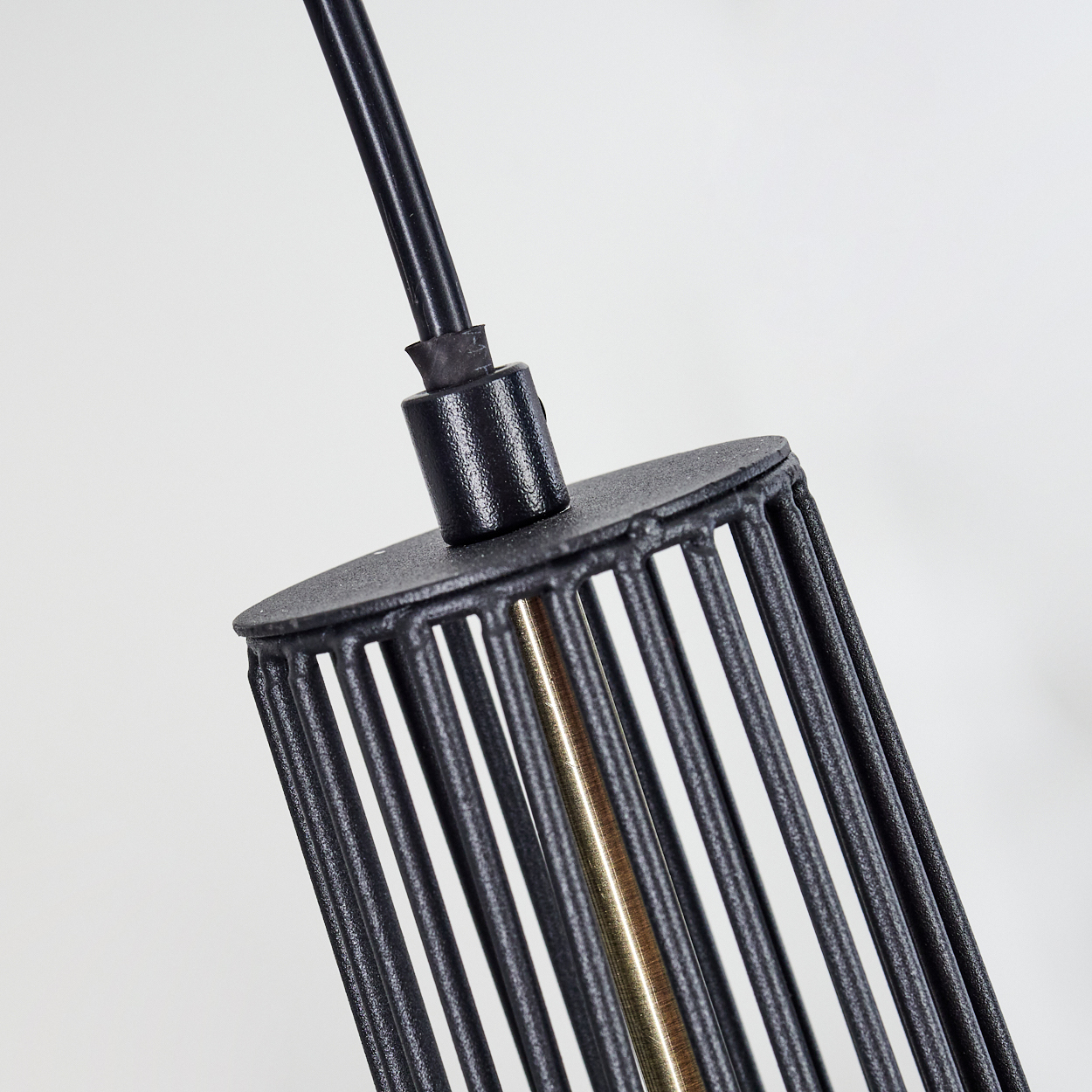 Nahaufnahme eines schwarzen Metalllampenschirms mit vertikalen Stäben und einem messingfarbenen Mittelteil. Ein schwarzes Kabel erstreckt sich nach oben, vor weißem Hintergrund.