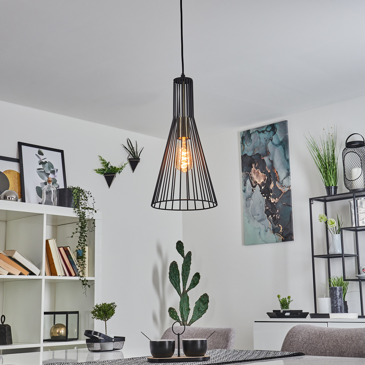 Ein modernes Esszimmer mit schwarzer Hängelampe, Kunstwerken, Regalen mit Dekoration, einem Kaktus und einem Esstisch mit Schalen.