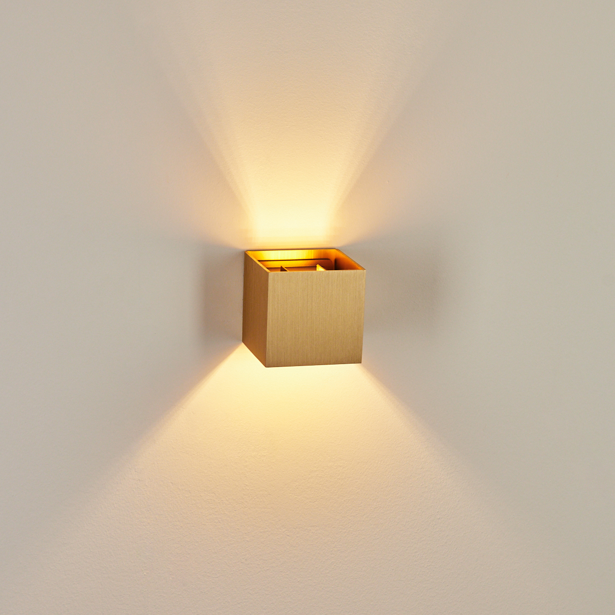Eine quadratische, goldfarbene Wandleuchte mit Licht, das an einer weißen Wand nach oben und unten scheint.