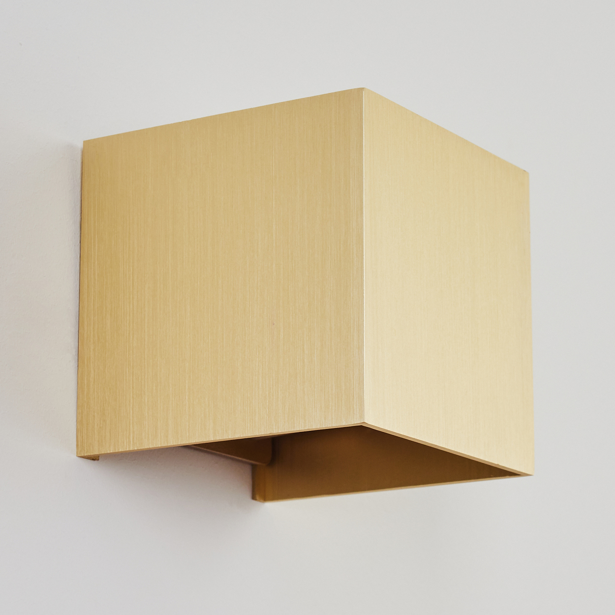 Eine goldene, geometrische Wandleuchte. Die Leuchte ist ein Würfel mit gebürsteter Metallstruktur, an einer weißen Wand befestigt.