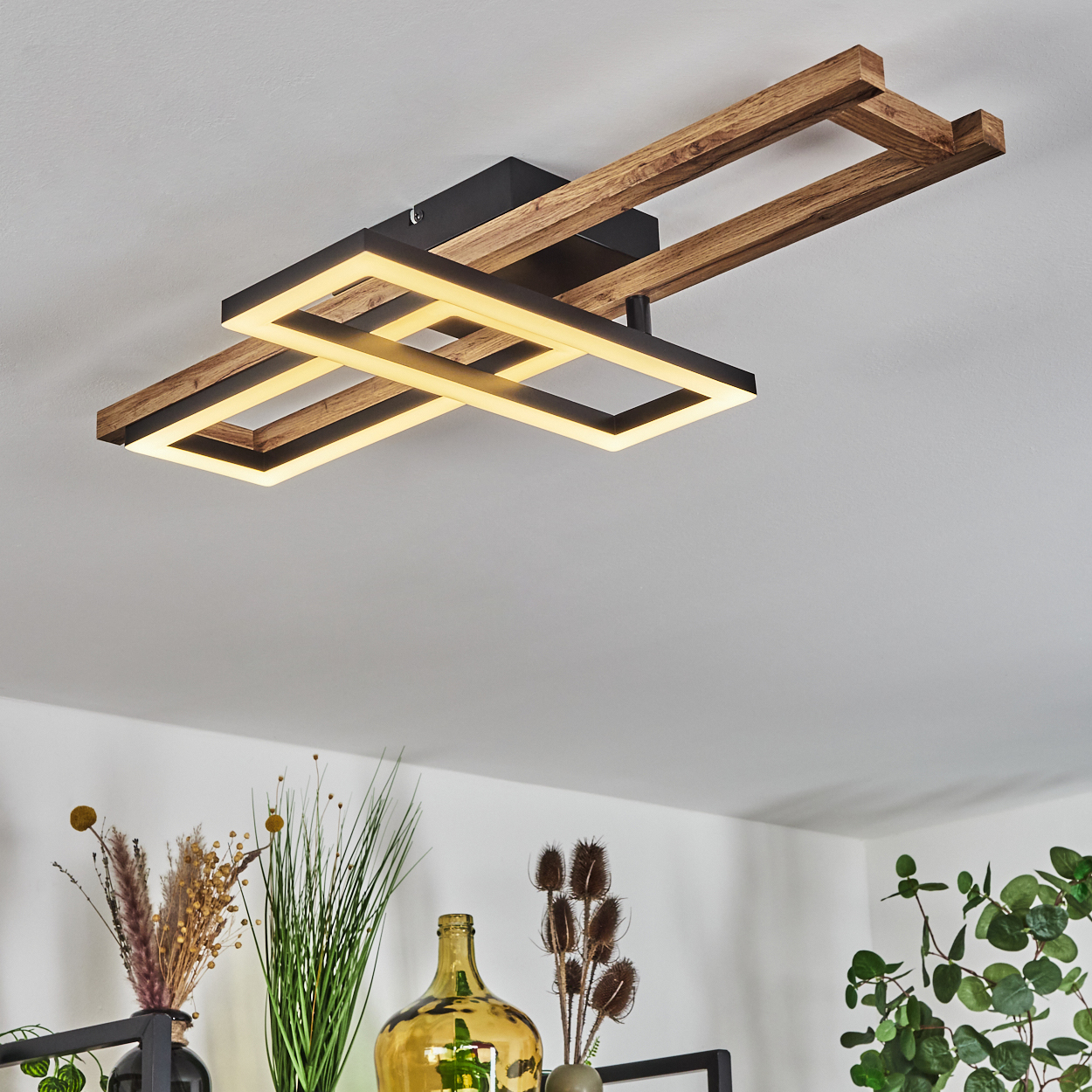 Eine moderne Deckenlampe mit Holzrahmen und schwarzem Sockel. Darunter dekorieren Vasen und Pflanzen den Raum.