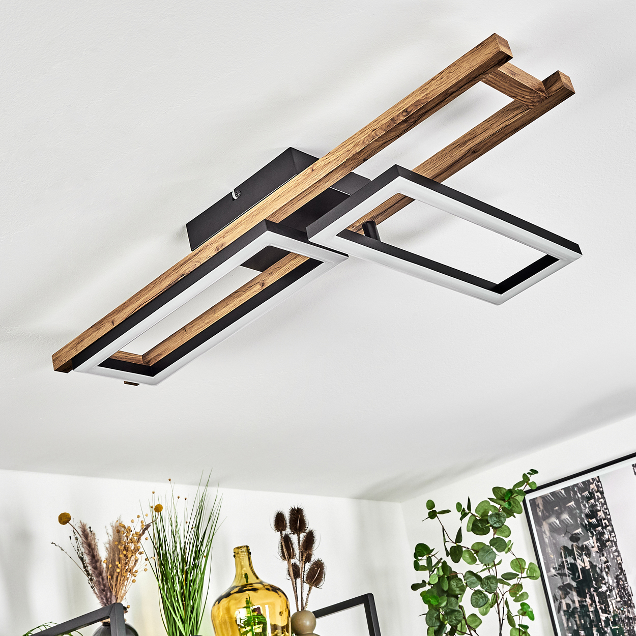 Eine moderne Deckenleuchte mit Holz- und schwarzen Rahmen. Darunter Dekoration und Pflanzen auf einem Regal. Weiße Decke und Wand.
