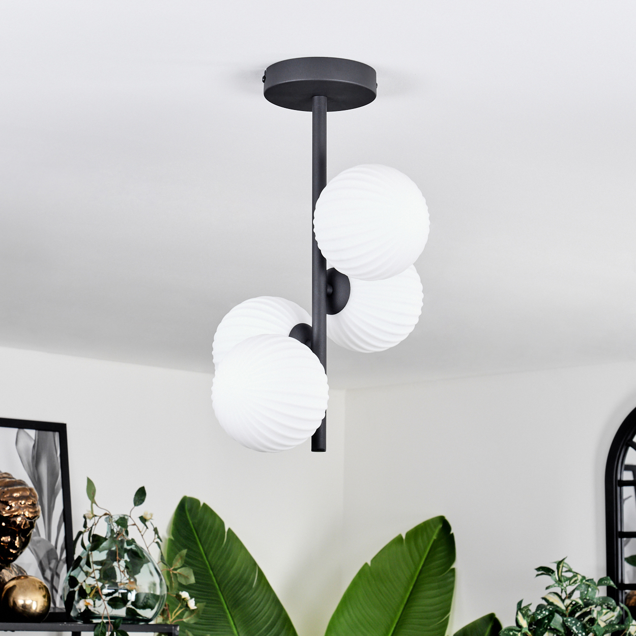 Eine moderne schwarze Deckenleuchte mit vier weißen, kugelförmigen Lampen. Grüne Pflanzen im Vordergrund.