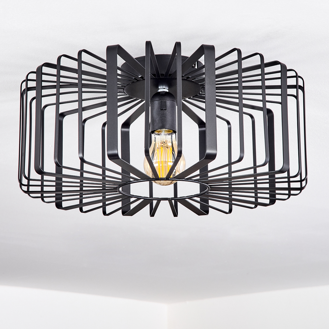 Eine schwarze Deckenleuchte im Käfigstil mit sichtbarer Glühbirne hängt vor einem weißen Hintergrund. Das Licht ist geometrisch und modern gestaltet.