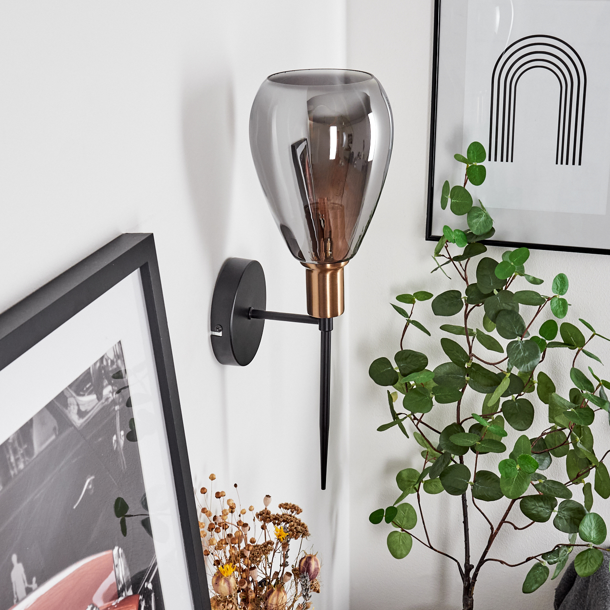 Eine Wandlampe mit grauem Glasschirm und schwarzem Metallarm. Ein gerahmtes Bild und eine Pflanze daneben.