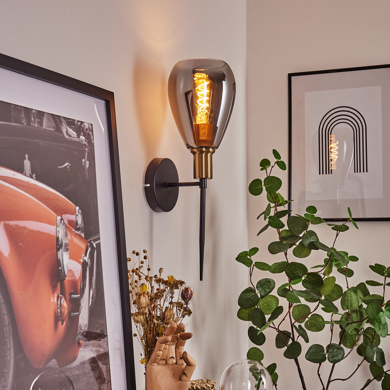 Eine Wandlampe mit rauchfarbenem Glasschirm und sichtbarem Glühfaden. Ein gerahmtes Bild eines Autos und Pflanzen.