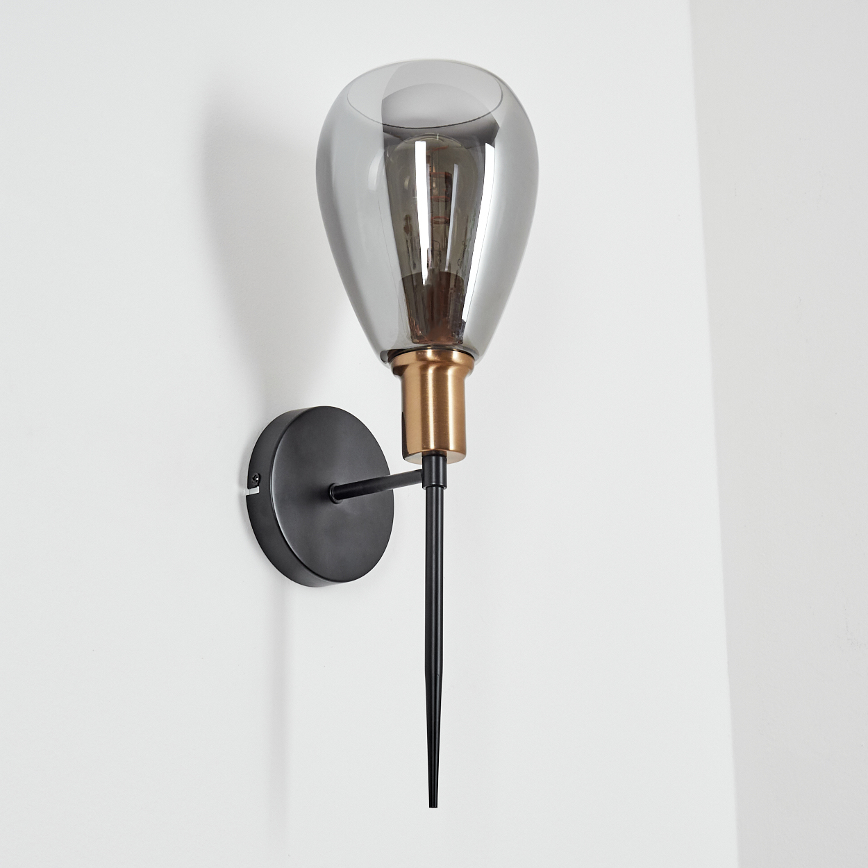 Eine Wandlampe mit einem tropfenförmigen Glasschirm. Sie hat einen schwarzen Arm und Sockel mit einem goldfarbenen Hals, vor einer weißen Wand.