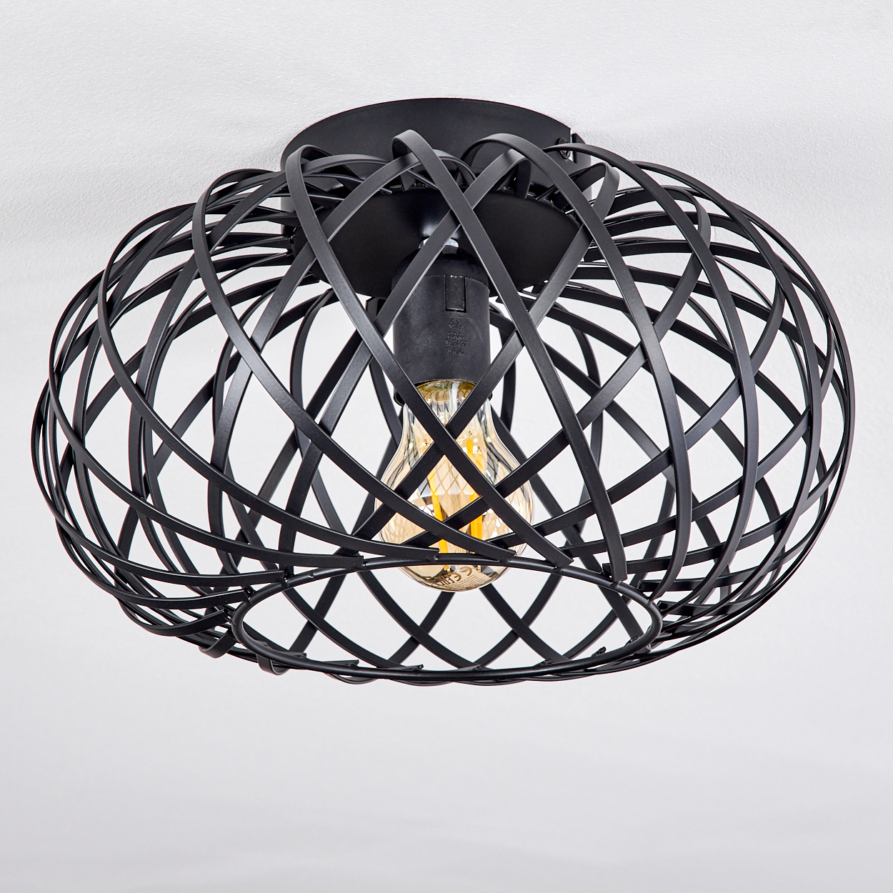 Eine schwarze, käfigartige Deckenlampe mit sichtbarer Glühbirne vor einer weißen Decke. Die Lampe hat ein geometrisches Design.