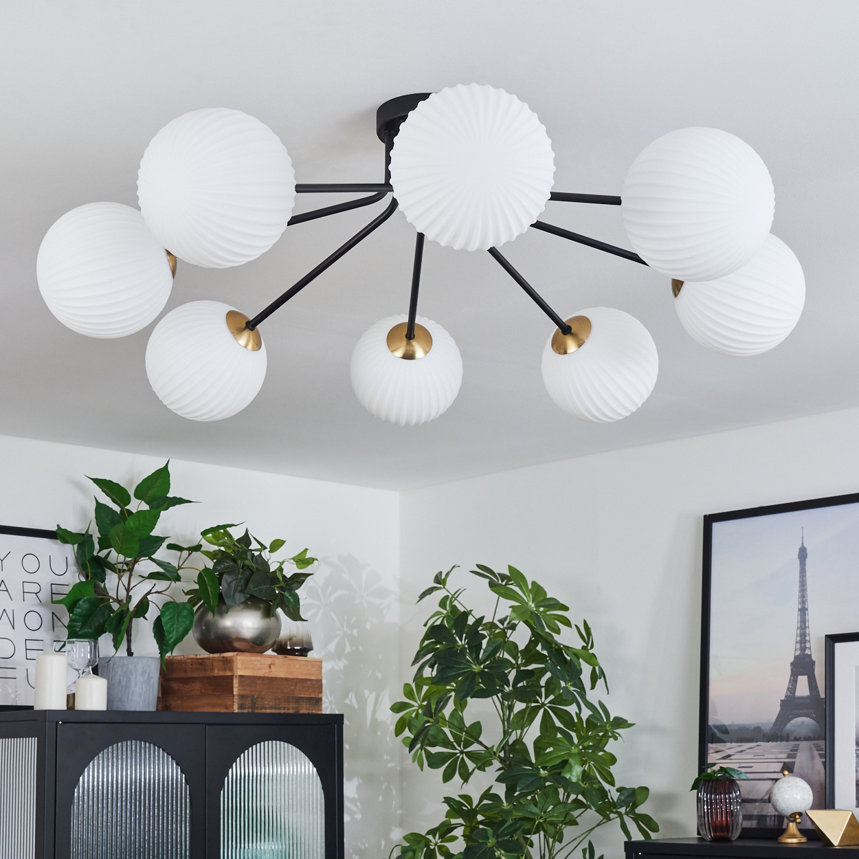 Moderne Deckenlampe mit weißen Kugelleuchten. Darunter ein schwarzer Schrank mit Pflanzen und einem gerahmten Eiffelturm-Druck.