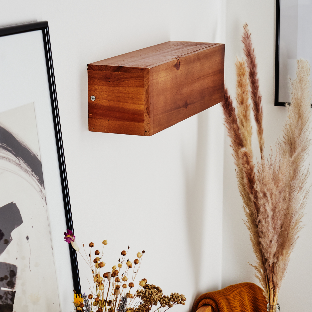 Ein Holzregal hängt an einer weißen Wand. Getrocknete Blumen und Pampasgras stehen in der Ecke. Links ist ein gerahmtes Bild.