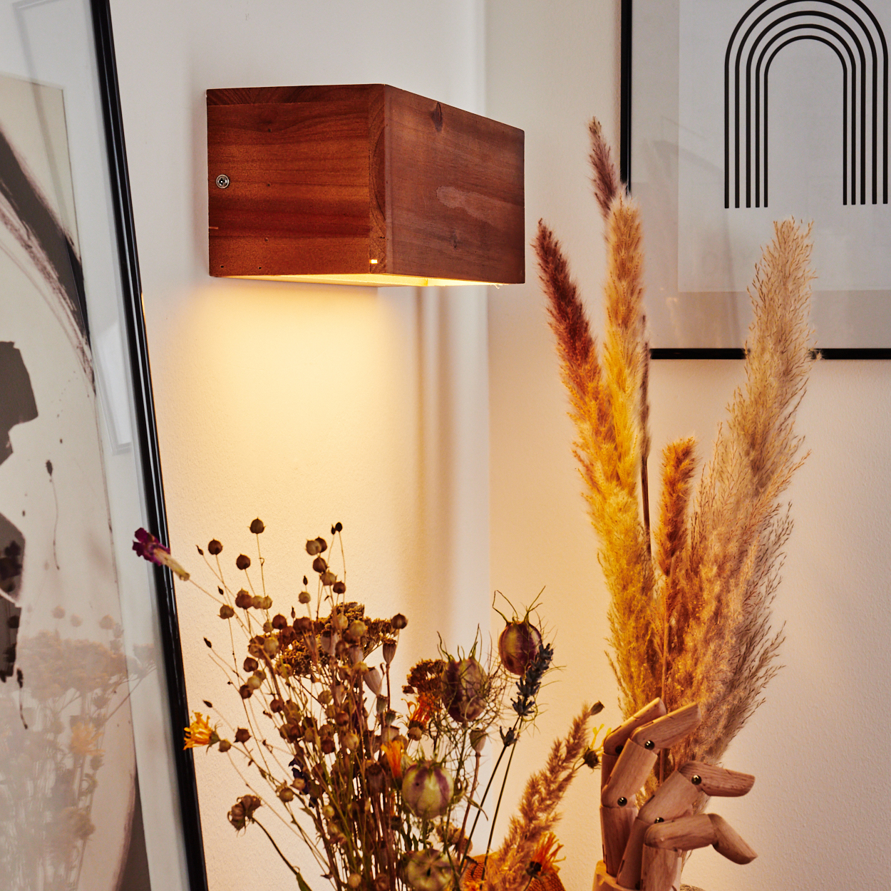 Holzlampe an der Wand mit getrockneten Blumen und Kunstwerken. Warmes Licht beleuchtet den Bereich. Dekorative Elemente im Vordergrund.