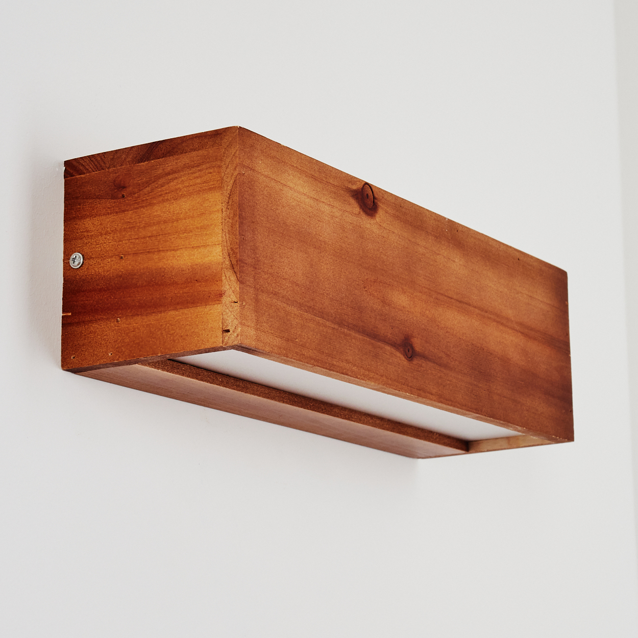 Ein rechteckiger Holzkasten ist an einer weißen Wand befestigt. Die Box ist braun und hat einen weißen Boden, der von innen beleuchtet wird.