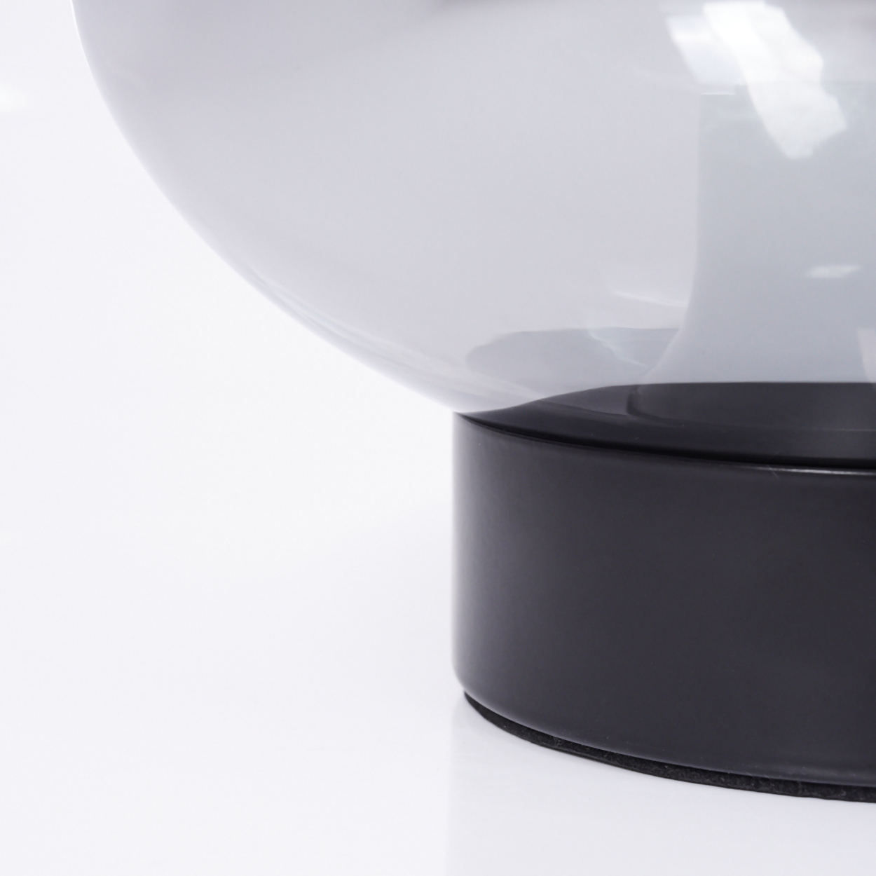Nahaufnahme einer Lampe. Ein klarer, runder Glasschirm sitzt auf einem schwarzen, zylindrischen Sockel. Weißer Hintergrund.