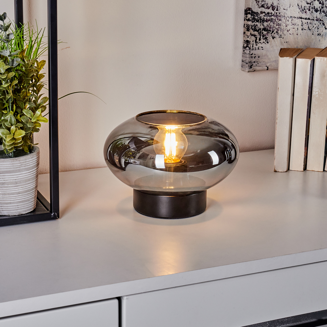 Eine moderne Lampe aus Rauchglas mit einer brennenden Glühbirne steht auf einer weißen Oberfläche. Ein Pflanze und Bücher sind im Hintergrund.